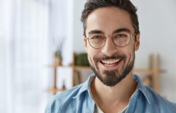 Close up shot of cheerful satisfied attractive male with stubble, has broad smile, wears round spectacles, rejoices success at work, stands against cozy interior. Fashionable designer glad be praised