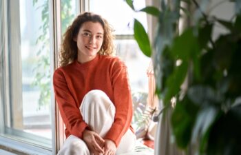 Pretty serene young woman sitting on windowsill relaxing at home looking at camera. Smiling calm lady chilling in apartment, dreaming, thinking of peaceful time enjoying peace of mind. Portrait