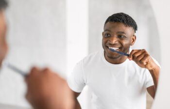 Handsome African American millennial guy brushing teeth with toothbrush stands by the mirror, caring for his oral health in modern bathroom. Daily dental care routine. Selective focus
