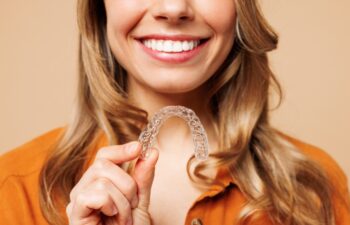 Close up cropped young happy woman wear orange shirt casual clothes hold in hand invisible transparent aligners, invisalign bracer isolated on plain pastel light beige background. Lifestyle concept